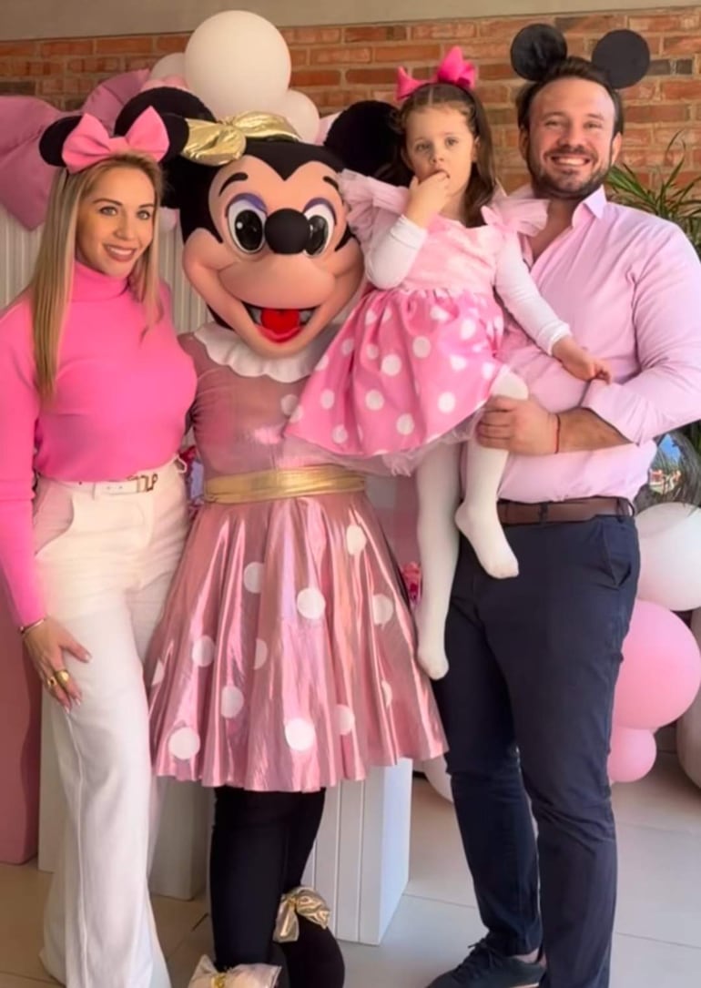 Mujer disfrazada de Minnie Mouse sonríe entre un hombre con camisa rosa y una niña pensativa en vestido rosa con puntos blancos.