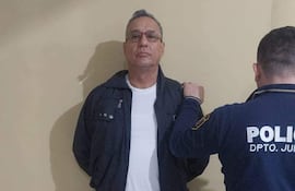 Hugo Javier González esposado en camiseta blanca y abrigo oscuro, con expresión tranquila, rodeado por un policía con uniforme.
