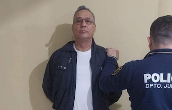 Hugo Javier González esposado en camiseta blanca y abrigo oscuro, con expresión tranquila, rodeado por un policía con uniforme.
