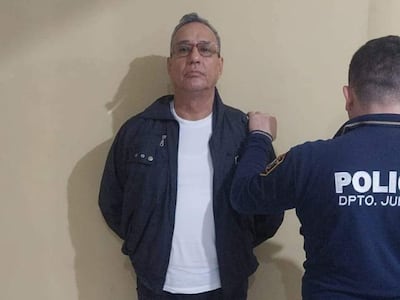 Hugo Javier González esposado en camiseta blanca y abrigo oscuro, con expresión tranquila, rodeado por un policía con uniforme.