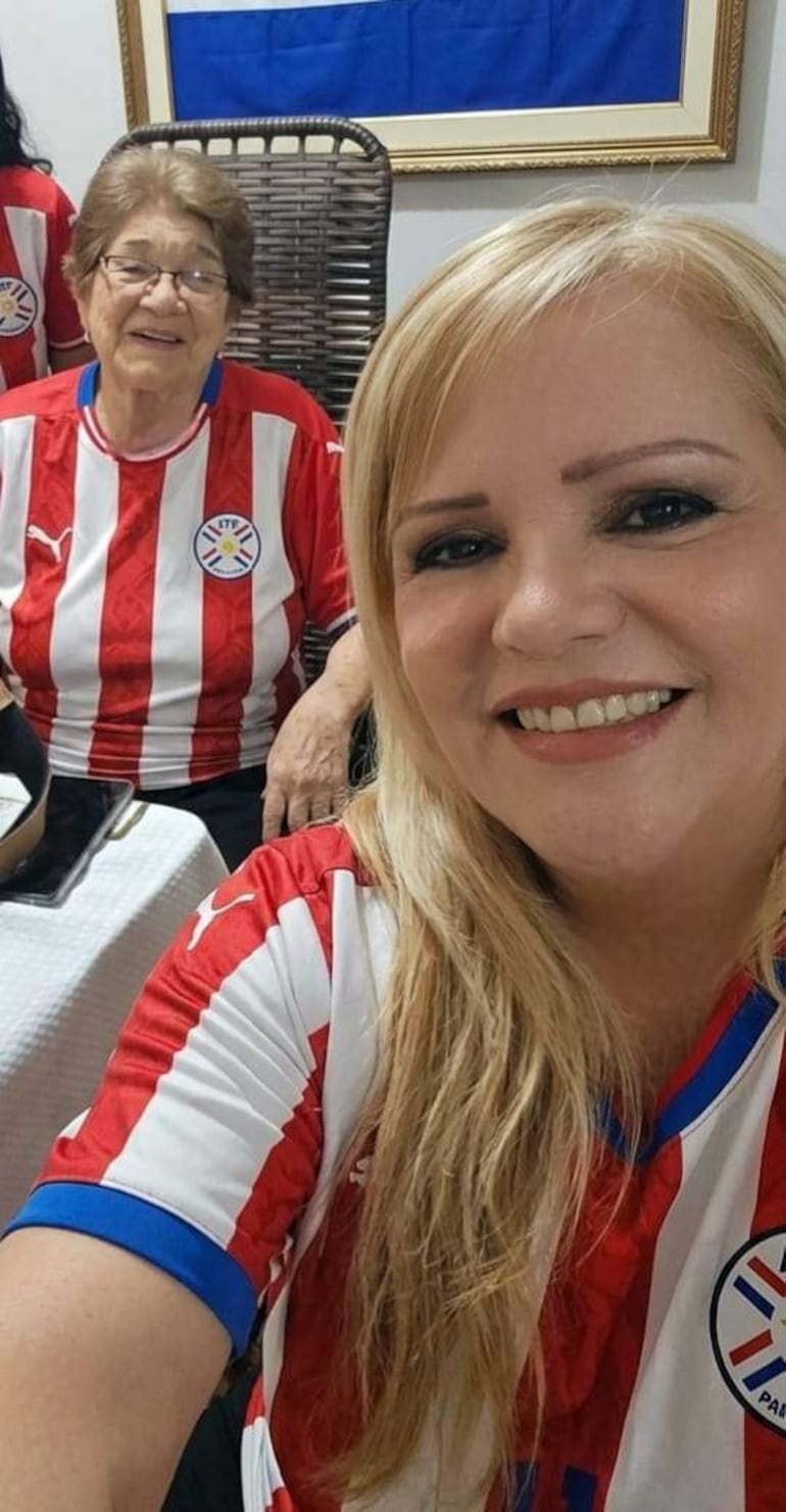 Dos mujeres sonríen, una en primer plano y otra sentada, con camisetas rojas y blancas de la selección paraguaya. Decoración patriótica en el fondo.