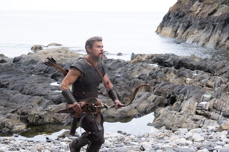 Hombre de mediana edad con barba y armadura de cuero, sosteniendo un arco, camina por rocas junto al mar.