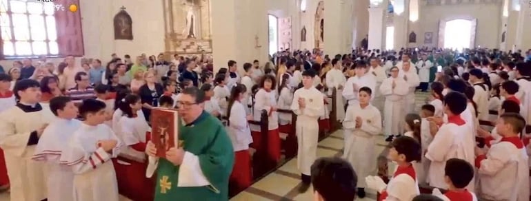 Grupo de monaguillos con ropas blancas y sobrehombros rojos, atentos a un sacerdote con túnica verde durante ceremonia en la catedral.