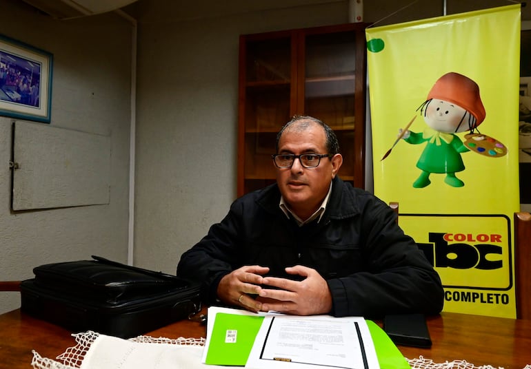 Hombre con gafas y chaqueta oscura hablando en una mesa, rodeado de documentos y un maletín negro.