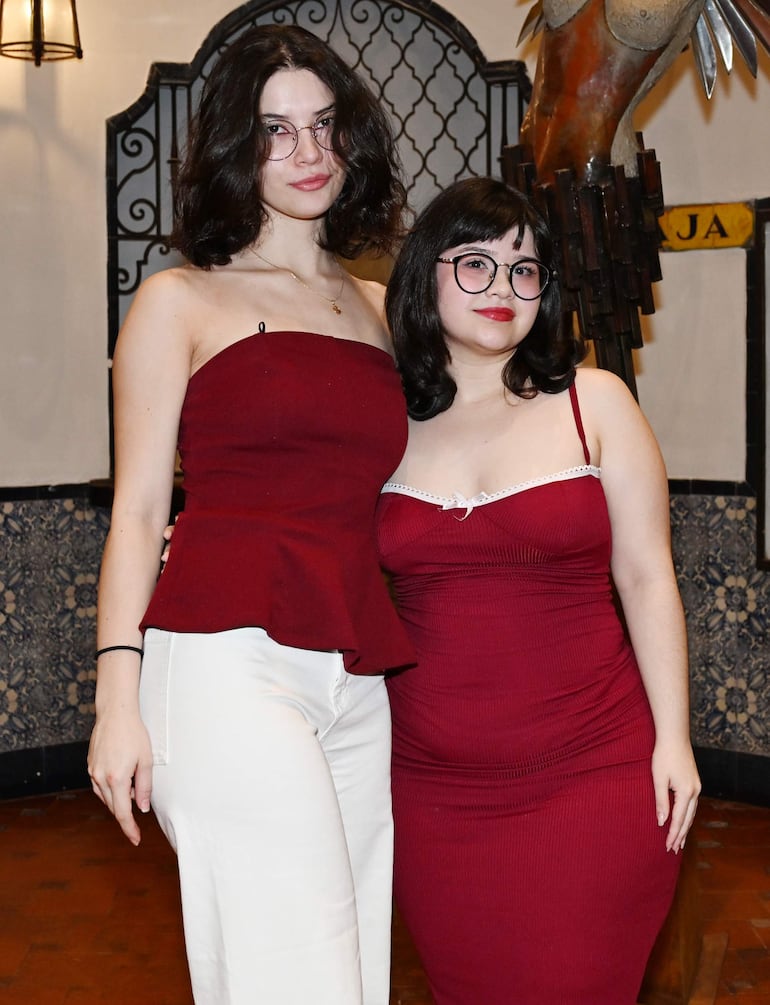 Mujeres sonrientes, una con blusa burdeos y pantalones blancos, otra en vestido burdeos, rodeadas de arte en un interior decorativo.