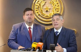 Patrick Kemper y Luis Ramírez en conferencia de prensa, ambos con trajes azules, micrófonos visibles y emblema gubernamental al fondo.