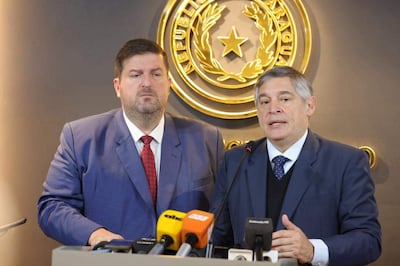 Patrick Kemper y Luis Ramírez en conferencia de prensa, ambos con trajes azules, micrófonos visibles y emblema gubernamental al fondo.