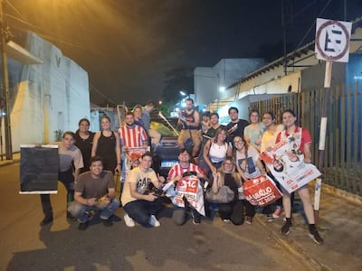 Veinte jóvenes sonrientes en una calle, algunos sentados en el suelo, sosteniendo carteles y visiblemente felices.