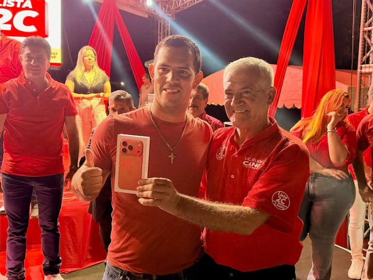 Dos hombres en camisetas rojas posan sonrientes; uno sostiene un teléfono y muestra un gesto de aprobación en un ambiente decorado con telas rojas.