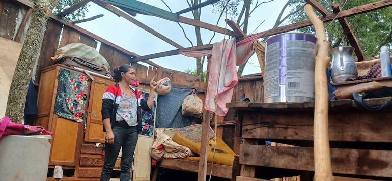 Mujer de pie en hogar improvisado, con balón de fútbol y señalando, en un ambiente precario de madera y textiles.