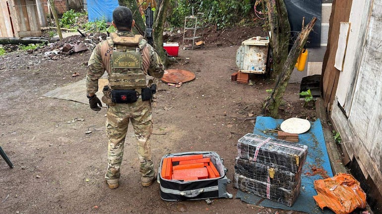 Agente de Policía Federal con uniforme camuflado de espaldas, inspecciona maletas en un ambiente rural y lluvioso.