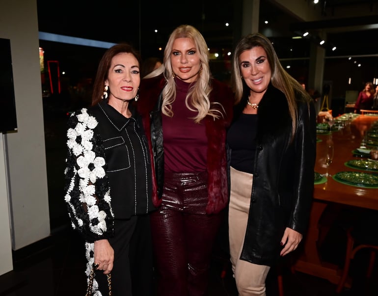 Tres mujeres sonríen, una con blusa negra, otra con abrigo rojo y la última en abrigo negro, en ambiente festivo.