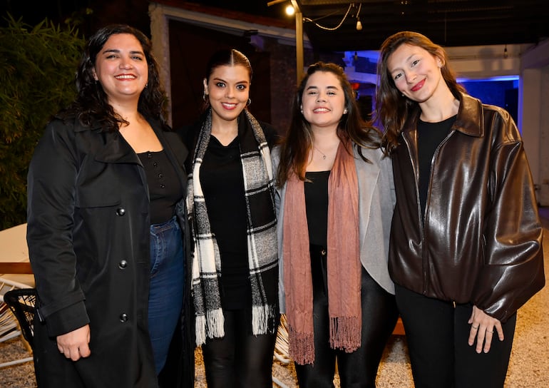 Cuatro mujeres posan juntas, luciendo atuendos oscuros y formales, en un ambiente de celebración iluminado.