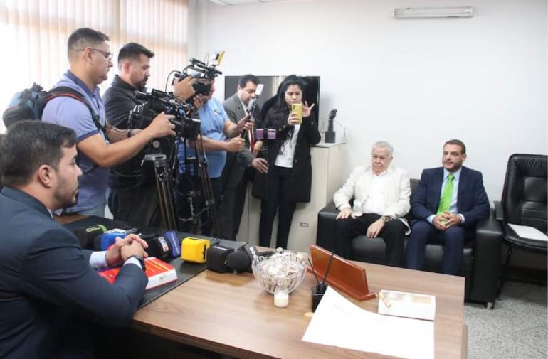 Ambiente de oficina con Jorge Brítez, hombre mayor en traje azul, rodeado de varios reporteros y cámaras, todos observando atentos.