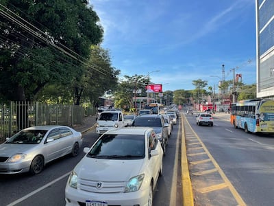 Escena urbana con tráfico, árboles a la izquierda y edificios a la derecha, sin personas visibles, durante el día.