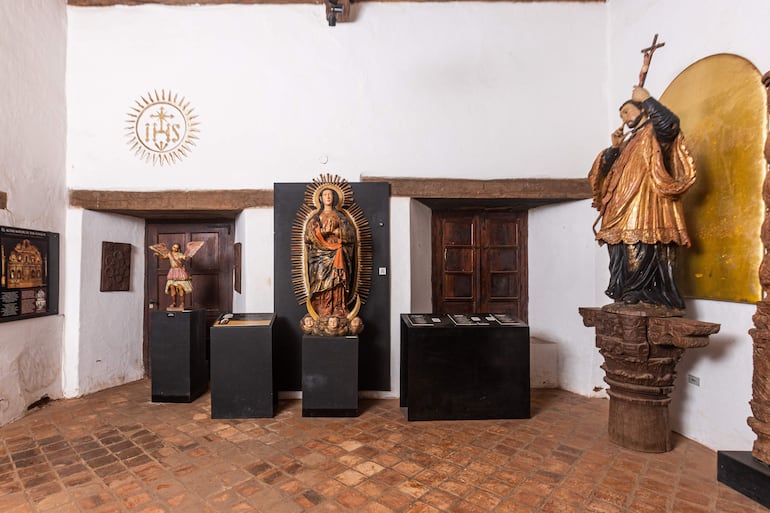 Virgen María con halo dorado, ángel colorido y santo con crucifijo, en un museo con paredes blancas y ambiente reverente.