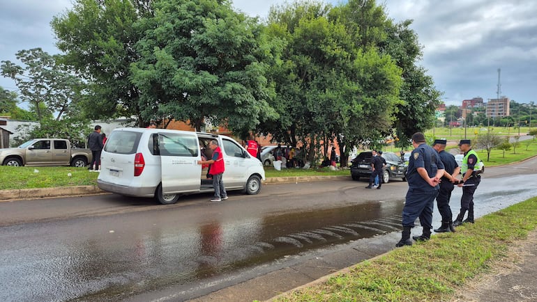 Hombre de camiseta oscura y pantalones claros junto a una furgoneta blanca, mientras policías observan en un entorno urbano con vegetación.