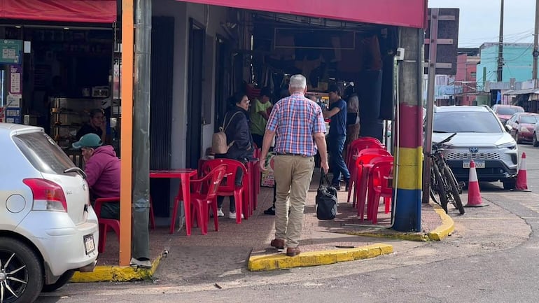 Hombre con gorra y sudadera rosa sentado, otros tres adultos conversan en un establecimiento comercial con mesas rojas.