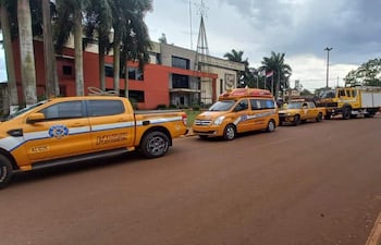 Filas de vehículos amarillos de emergencia y servicios públicos en una calle. Edificio moderno y banderas al fondo, cielo parcialmente nublado.