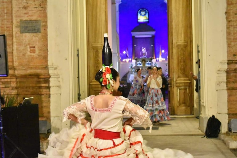 Mujer con vestido rojo y blanco baila en el centro, con una botella en la cabeza, rodeada de bailarinas en un ambiente festivo.