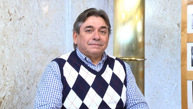Ing. Reinaldo Delgado, hombre mayor con chaleco de rombos y camisa de cuadros, sentado en oficina con fondo de mármol.