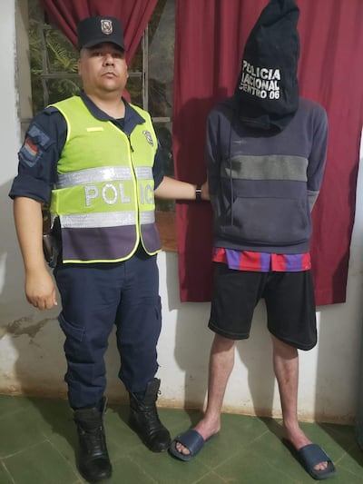 Agente de policía en uniforme serio junto a Iván Matías Aguada Araujo, cubierto con bolsa negra y en sudadera gris, en ambiente de vivienda.