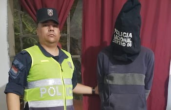 Agente de policía en uniforme serio junto a Iván Matías Aguada Araujo, cubierto con bolsa negra y en sudadera gris, en ambiente de vivienda.