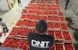Hombres organizan cajas de tomates en un almacén cerrado, uno observa desde una estructura elevada, todos con expresión de concentración.