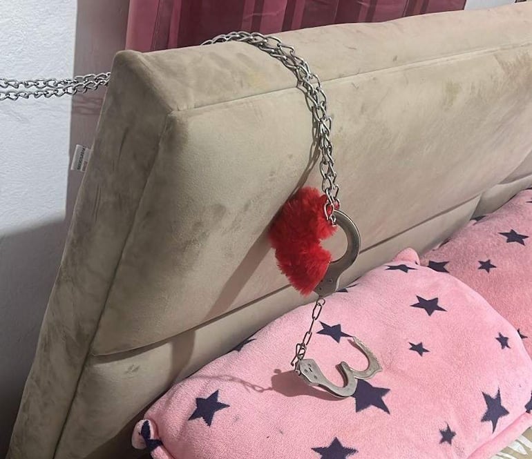 Cabecero de cama con cadena y esposas, decorado con almohadas rosas con estrellas azules y un peluche rojo.