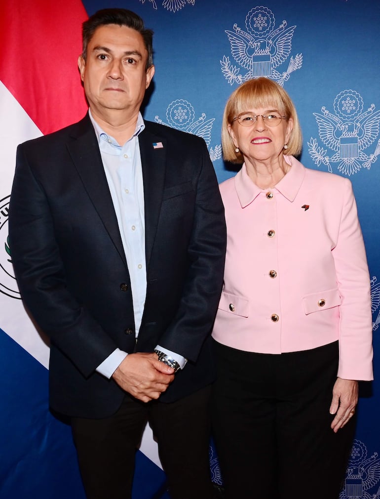 Mujer con blusa rosa y gafas junto a hombre con blazer oscuro en evento diplomático con símbolos institucionales de Paraguay.