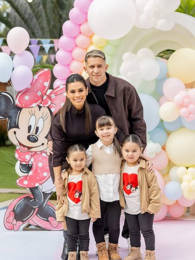 Jorge Morel sonríe junto a una mujer en atuendo oscuro y tres niñas con camisetas de Disney, rodeados de globos coloridos.