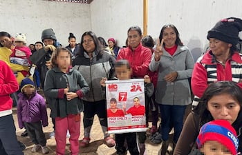 Emílce Vera Díaz en el centro, con cartel de campaña, rodeada de personas en un ambiente comunitario informal, vestidas con abrigos.