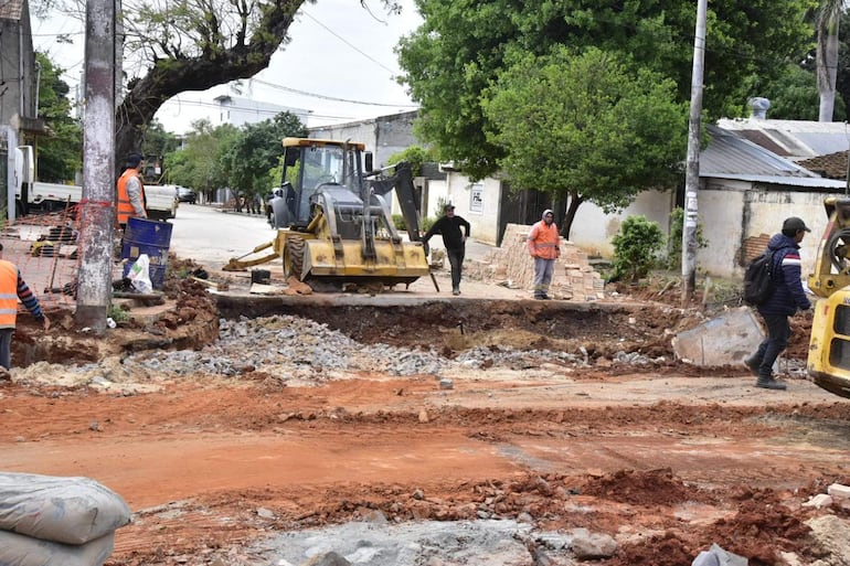 Cuatro trabajadores en ropa de trabajo supervisan obras en calle, incluyendo a un operario en retroexcavadora amarilla.