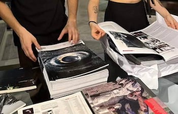 Hombre en camiseta negra sostiene revistas mientras una mujer a su lado revisa documentos en un entorno artístico.