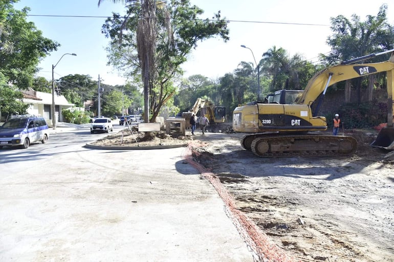 Maquinaria pesada Caterpillar en obra vial, rodeada de árboles, con vehículos en movimiento y trabajadores en chalecos naranjas.