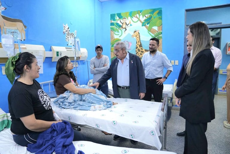 Isaías Fretes en chaqueta oscura interactúa con mujer en camiseta marrón, mientras otras personas observan en un hospital infantil decorado.