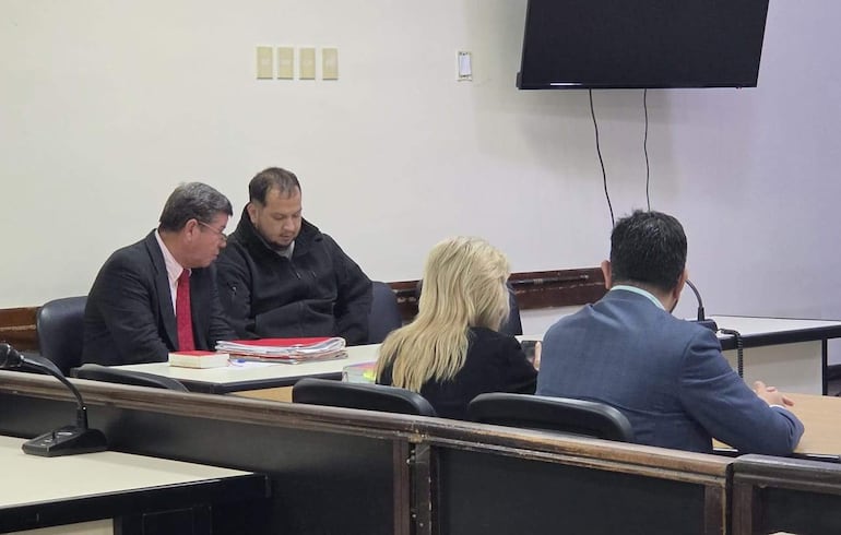 Interacción entre hombre de traje negro y corbata roja, mujer rubia y hombre de chaqueta azul claro en un ambiente tribunal.
