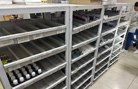 Mujer de pie con blusa blanca y pantalones oscuros en farmacia con estanterías vacías, mostrando un ambiente de desabastecimiento.