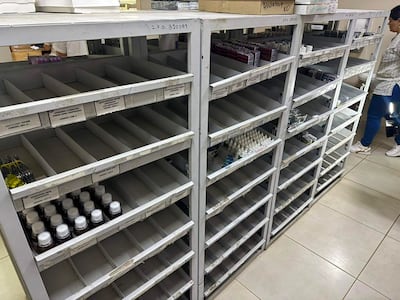 Mujer de pie con blusa blanca y pantalones oscuros en farmacia con estanterías vacías, mostrando un ambiente de desabastecimiento.