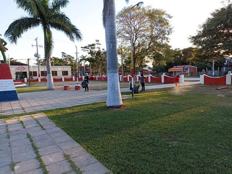 Dos personas en movimiento: una barre con abrigo rojo y otra recoge residuos con bolsa negra en un parque con césped y palmeras.