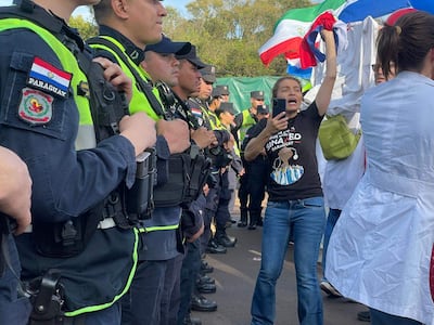 Mujer con camiseta de SINADES levanta la mano con un teléfono mientras policías alineados observan en un entorno cargado de banderas paraguayas.