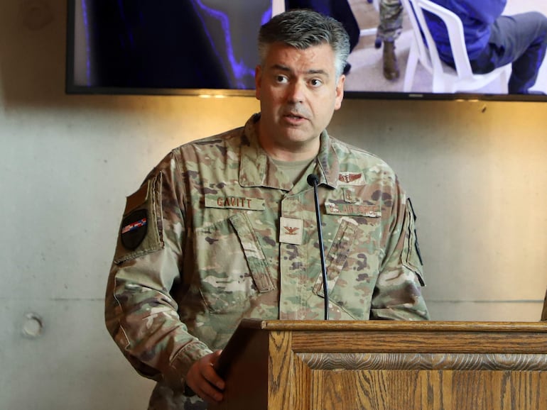 Gavitt, en uniforme militar, habla frente a un podio en un salón de conferencias.