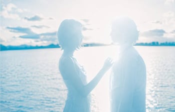 Mujer de cabello corto y prenda clara junto a hombre con camisa blanca, ambos en un ambiente luminoso y etéreo frente a agua y cielo.