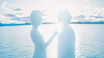 Mujer de cabello corto y prenda clara junto a hombre con camisa blanca, ambos en un ambiente luminoso y etéreo frente a agua y cielo.