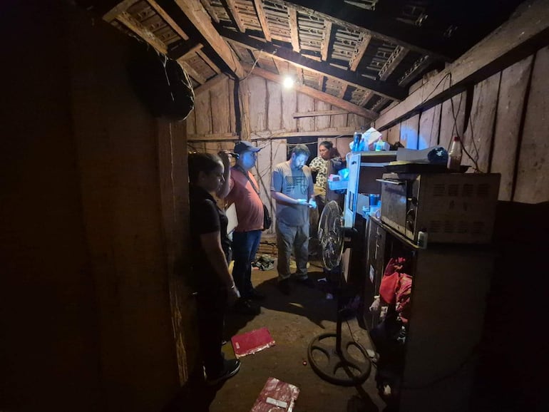 Hombres manejan objetos en un interior rústico, con un ventilador visible y luz tenue. Una mujer observa de pie apoyada en la pared.
