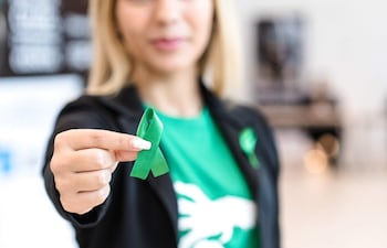 Mujer con camiseta verde y abrigo negro sostiene un lazo verde en un ambiente interior neutro.