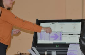 Mujer con blusa naranja y gafas interactúa con máquina de votación en ambiente electoral iluminado y despejado.