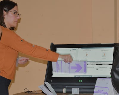 Mujer con blusa naranja y gafas interactúa con máquina de votación en ambiente electoral iluminado y despejado.