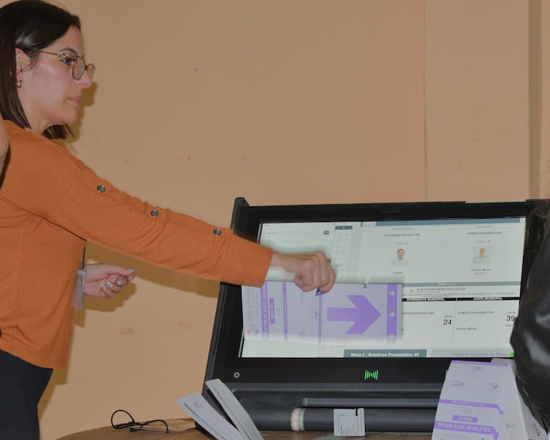 Mujer con blusa naranja y gafas interactúa con máquina de votación en ambiente electoral iluminado y despejado.