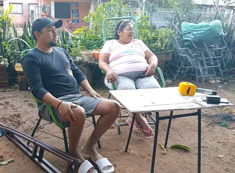 Juan Carlos Bogado, con camiseta negra, sentado en silla, escucha a su madre Delosantos Rodríguez, vestida con ropa estampada, en un entorno natural.